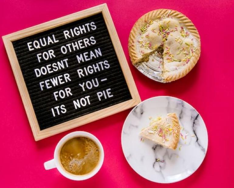 Image is a flat lay from above on a hot pink background. There's a pegboard in the top left showing "Equal rights for others does not mean fewer rights for you - it's not pie" in all caps. The pegboard has a wooden frame and black background with white pegs. In the top right is pie with a slice missing and sprinkles on top. In the bottom right is the slice of pie on a marbled plate. In the bottom left is a white mug filled with coffee.